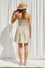 Woman wearing a beige dress and straw hat against a white curtain background