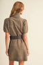 A woman shown from the back wearing a short-sleeve knit turtleneck dress in a taupe-brown color with a dark brown belt at the waist, set against a plain cream background.