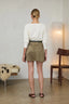 A woman is shown from behind wearing a white long-sleeve top and a brown suede mini skirt with a dark belt, standing in a minimalist room with a floor lamp and wooden chair.