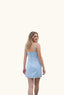 A woman with blonde hair stands outdoors in front of a large concrete stadium, wearing a light blue sleeveless mini dress and looking back over her shoulder.