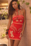 Woman wearing a red dress with white patterns leaning against a wall outdoors.