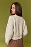 A woman with dark hair and glasses, shown from the back, wearing a cream-colored chunky knit sweater with cable detailing on the sleeves, set against a solid olive green background.