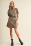 A woman models a short-sleeve knit mock-neck dress in a heathered taupe-brown color, styled with a dark brown belt and black ankle boots, against a solid cream background.
