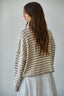A woman with long brown hair is shown from the back wearing a beige and white horizontally striped cropped knit sweater over a white tiered dress, set against a plain light gray wall.