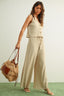 Woman in a beige sleeveless top and wide-leg pants holding a woven bag against a plain background
