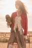 A woman stands outdoors in a sunlit field wearing a red cardigan, white lace blouse, and a brown-and-white plaid skirt, while holding a dark textured bag.