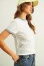Woman wearing a light gray ribbed t-shirt with short sleeves, blue jeans, and a beige cap on a plain background.