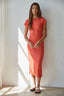 Woman wearing a coral dress standing against a white wall with sunlight casting shadows.