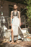 A woman stands outdoors in a rustic courtyard wearing a sleeveless bohemian-style maxi dress with a high slit. The dress has a cream base with dark brown geometric patterns and a draped V-neckline.