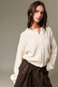 A woman wearing a cream-colored, textured waffle-knit button-up cardigan sweater and dark brown high-waisted pants, photographed against a neutral gray studio background.