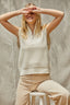 Woman wearing a beige sleeveless top and pants sitting on a stool against a neutral background