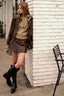 A woman stands against a white brick wall wearing a dark brown leather jacket, matching mini skirt, a beige knit sweater, and black knee-high boots.