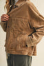 A cropped view of a person wearing a brown corduroy jacket with front flap pockets and dark gray jeans against a plain light beige background.