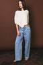 Woman wearing a white sweater and blue jeans against a brown background