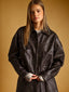 A woman wearing a dark brown faux-leather jacket with contrast stitching and a gray undershirt, standing against a solid warm brown background.