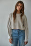 Woman wearing a beige ribbed sweater and blue jeans against a plain background