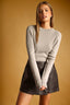 A woman modeling a light beige crewneck knit sweater paired with a dark gray wrap mini skirt, set against a solid warm tan background.
