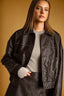 A woman wearing an oversized dark brown faux-leather jacket over a ribbed gray top, posed against a warm mustard-yellow studio background.