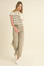 A woman modeling a short-sleeve knit top with beige and white horizontal stripes, paired with wide-leg beige trousers and white sneakers, against a solid cream background.