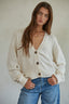 Woman wearing a cream cardigan with brown buttons and blue jeans against a plain background