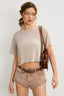 Woman wearing a beige crop top and shorts with a brown belt, holding a brown leather bag against a white background.