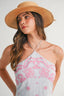Woman wearing a light blue halter dress with pink floral embroidery and a straw hat on a plain background