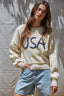Woman wearing a cream sweater with 'USA' text and denim shorts against a textured wall.