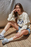 Woman wearing a cream sweater with 'USA' text, denim shorts, and white sneakers sitting on a wooden deck.