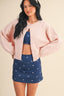 Woman wearing a pink cardigan, blue top, and blue skirt with white patterns on a beige background