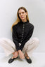 Woman wearing a black blouse with white buttons and beige pants against a white background