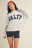 Woman wearing a gray sweatshirt with 'SALTY' text, holding it up to show the design.