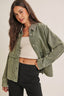 Woman wearing a green denim jacket over a white top and black pants on a plain background