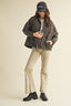 Woman wearing a dark jacket, cap, and beige pants on a beige background