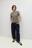 A woman modeling a taupe short-sleeve knit top with a decorative button detail down the front, paired with dark wide-leg trousers and black flats, standing against a plain white studio background.