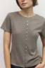 A woman wearing a short-sleeve crew-neck t-shirt in a heathered taupe gray color, featuring a vertical row of white buttons down the center front.