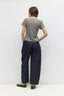 A woman shown from the back wearing a short-sleeve taupe-gray t-shirt, dark navy wide-leg trousers, and black flats, standing against a plain light gray studio background.