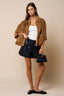 Woman wearing a brown jacket, white top, black shorts, and holding a black handbag against a beige background