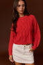 A woman wearing a bright red cable-knit cropped sweater and a white mini skirt, standing against a solid dark brown studio background.