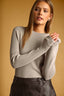 A woman wearing a light taupe ribbed knit long-sleeve sweater and a dark brown faux-leather skirt, posed against a solid warm tan background.