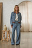 A woman stands in a rustic indoor setting wearing a light blue denim jacket and matching wide-leg jeans with brown contrast trim and side stripes. She has a white top underneath.