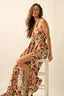 Woman wearing a rust-colored halter neck dress with leaf pattern sitting on a white stool against a plain background.