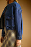 A person wearing a cropped blue denim jacket over a dark top and a beige plaid skirt, shown against a warm tan studio background.