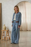 A woman stands in a styled indoor setting wearing a light blue denim jacket and matching wide-leg jeans with brown contrast trim. The room has textured walls, a wooden stool with a ceramic jug, and a cream curtain in the background.