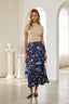 A woman modeling a beige short-sleeve mock neck top and a navy blue tiered midi skirt with a colorful floral print, standing in a bright, white-walled room with arched windows and columns.