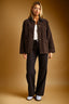 A woman stands against a warm tan studio background wearing a dark brown corduroy jacket over a white top, black wide-leg pants, and white sneakers.