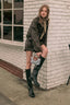 A woman posing outdoors against a white brick wall, wearing an oversized dark olive green jacket, a beige scarf, black knee-high boots, and holding a small gray handbag with brown leather trim.