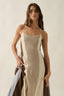 Woman wearing a beige ribbed dress holding a brown garment against a plain background