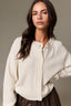 A woman wearing a cream-colored, textured knit cardigan with white buttons, paired with a dark brown skirt, against a neutral gray studio background.