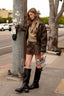 A woman stands on a city sidewalk wearing a brown faux-leather jacket, matching mini skirt, beige knit sweater, and black knee-high boots, while holding a small handbag near a utility pole.