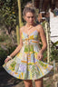 Woman wearing a colorful patterned dress in a natural setting with plants and a house.